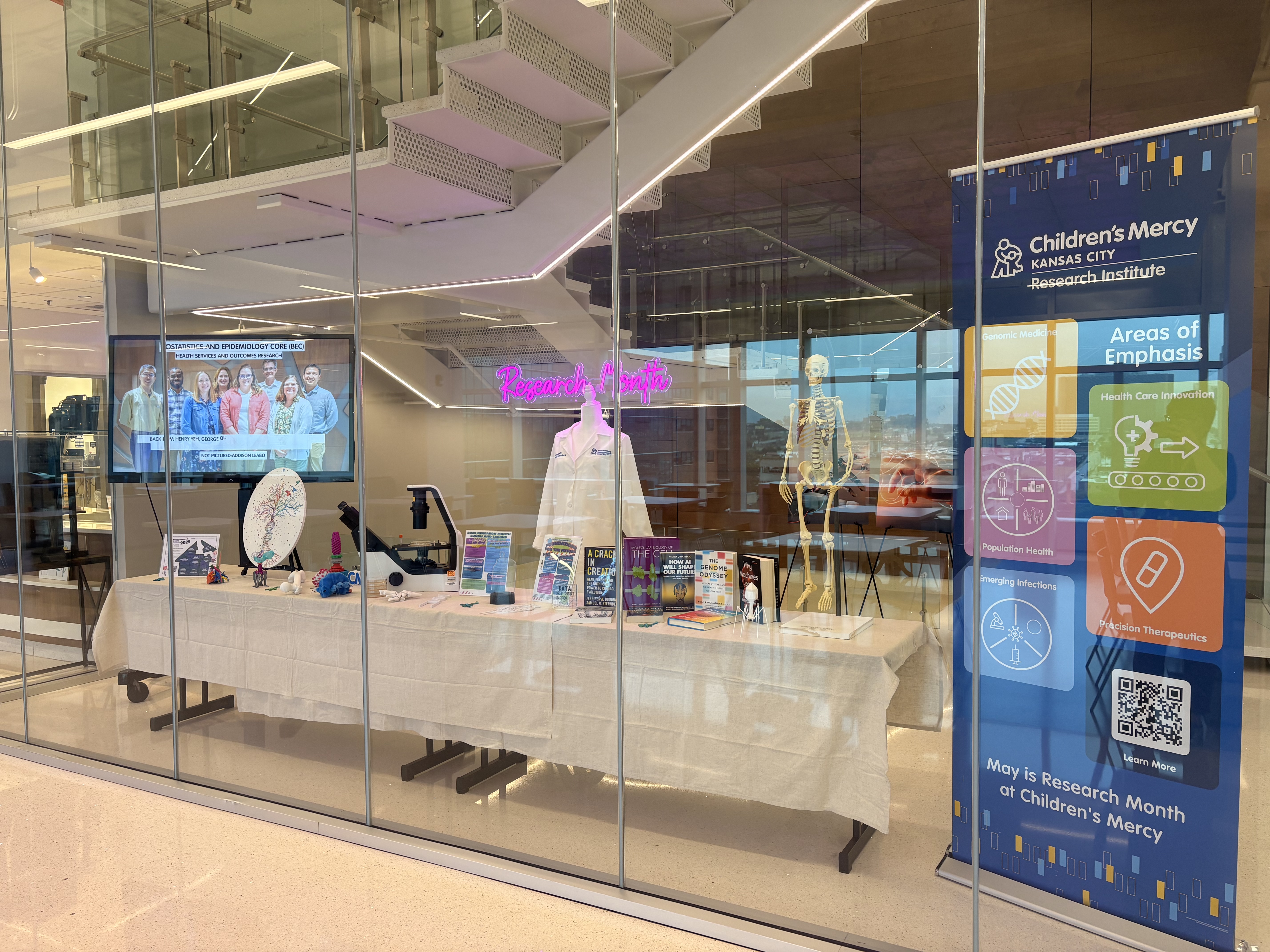 A display behind glass with a staircase behind it. The display is labeled Research Month. It contains a screen with photos rotating on it. A table with things on it. A mannequin wearing a lab coat and a skeleton.