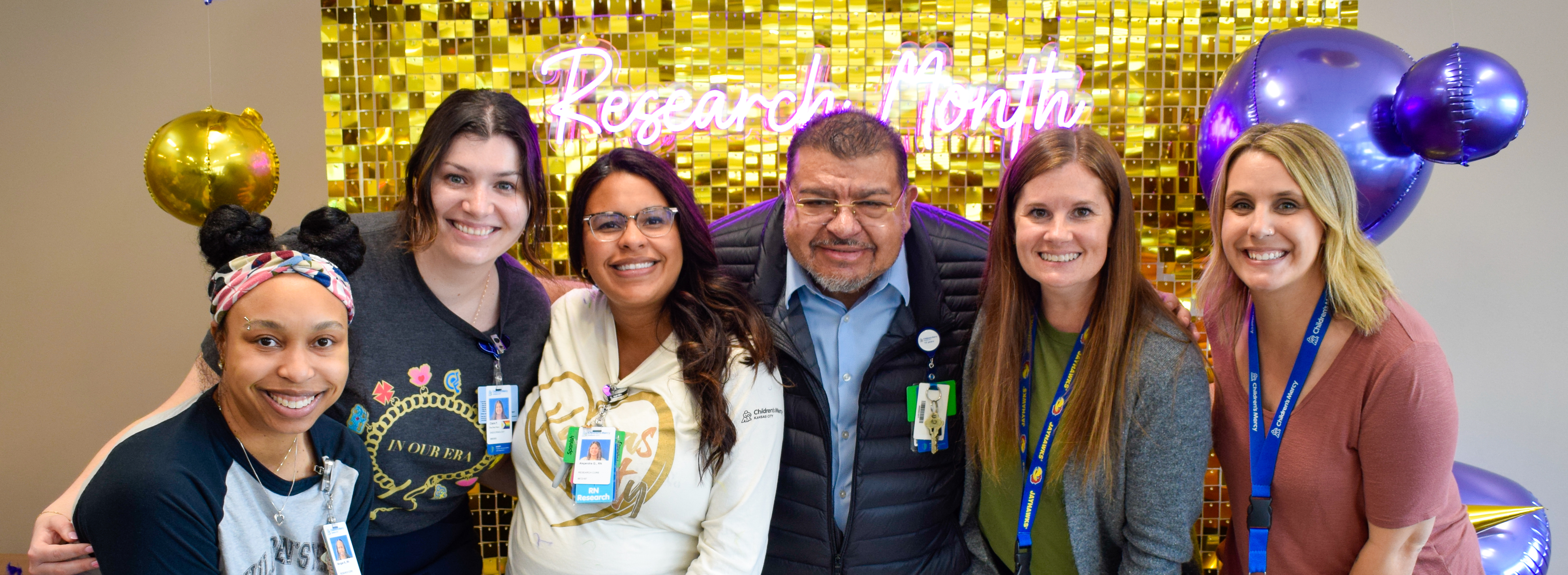 Six people, 5 of whom are women and 1 man, pose together, smiling. Behind them is a sign that says Research Month and balloons.