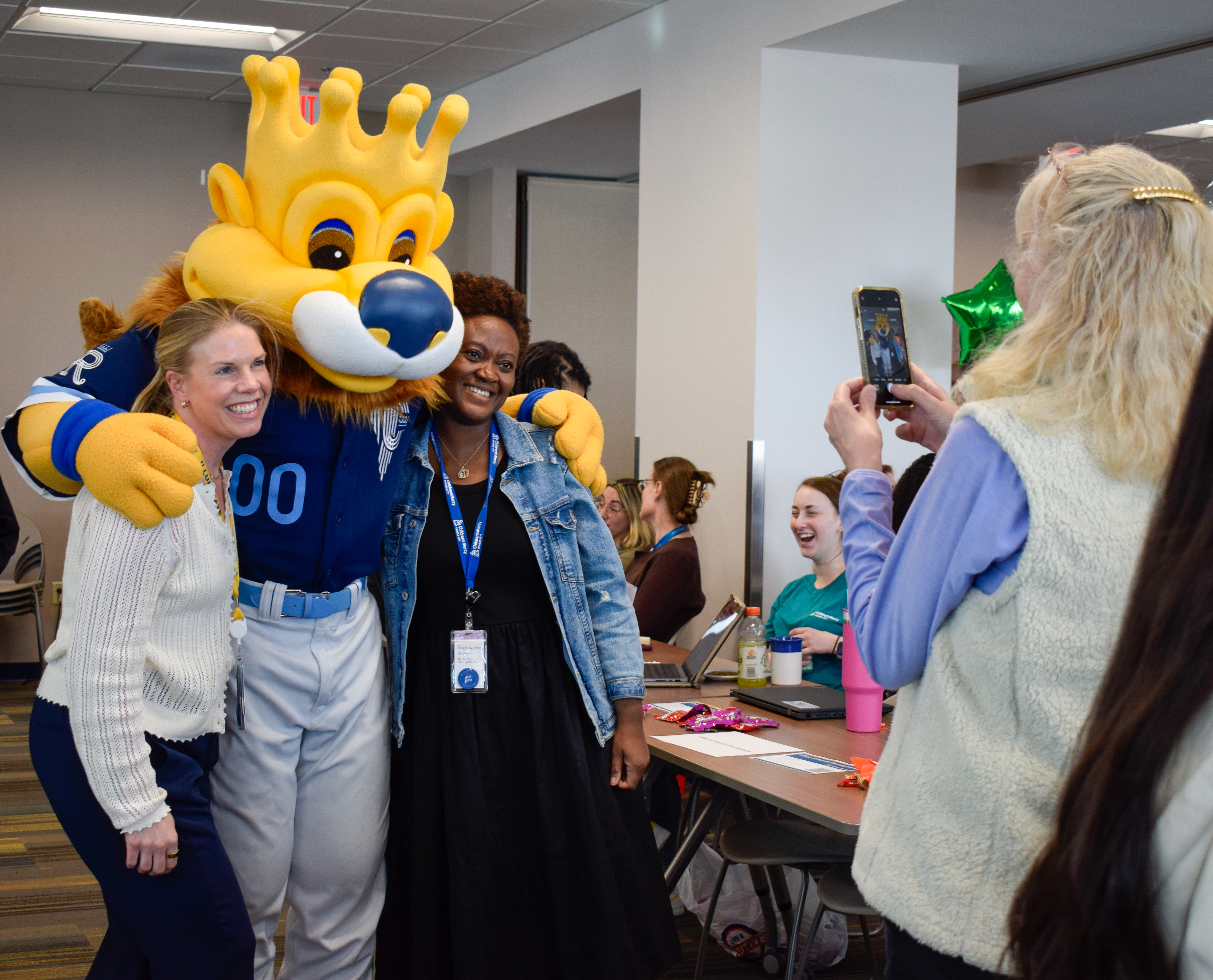 A mascot that looks like a lion and is wearing a baseball uniform poses with two women while another woman takes their photo on her cell phone.