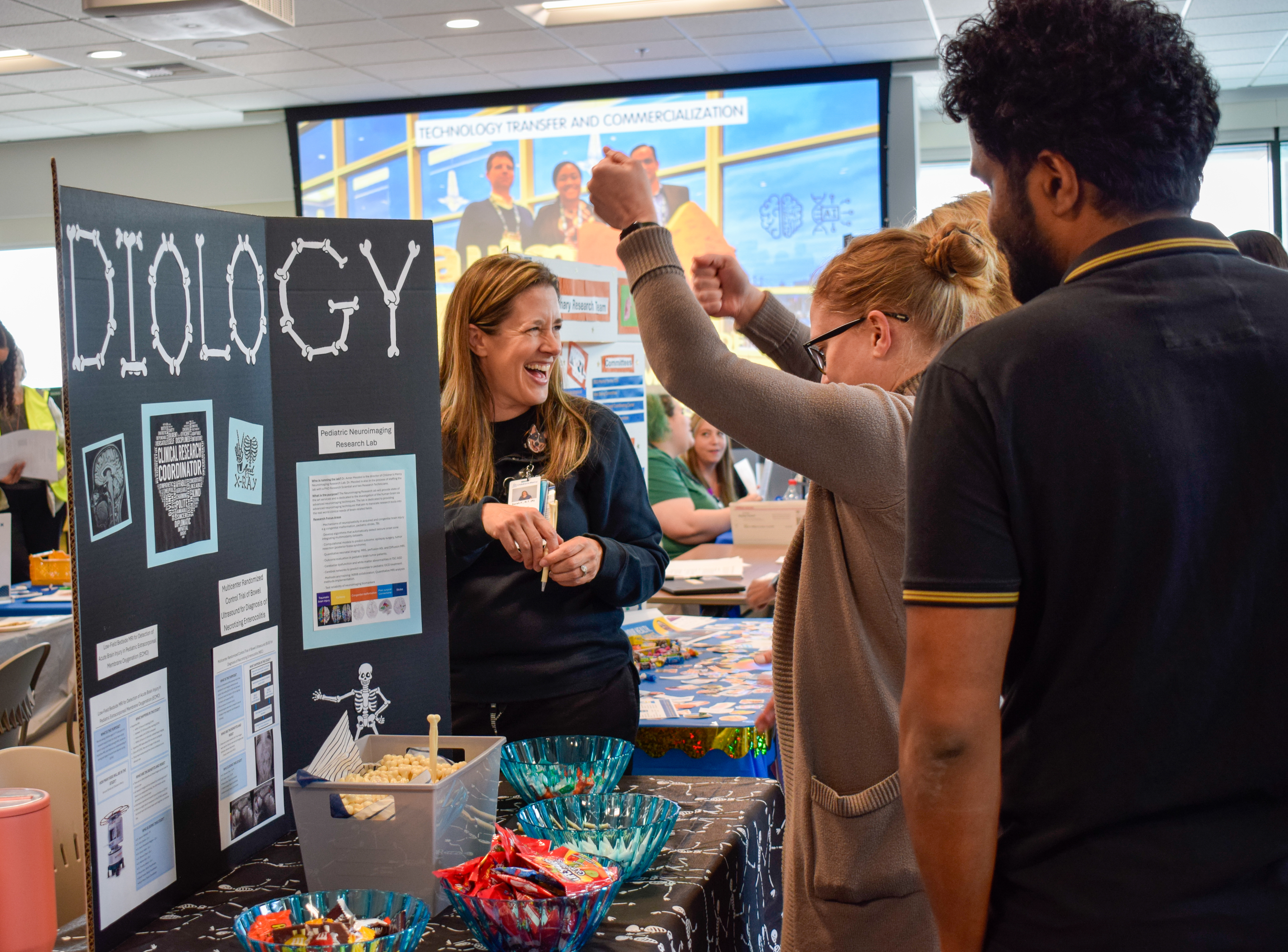 A woman stands at a table with a Radiology display, Another woman raises her hands in the air as she celebrates. A man looks on.