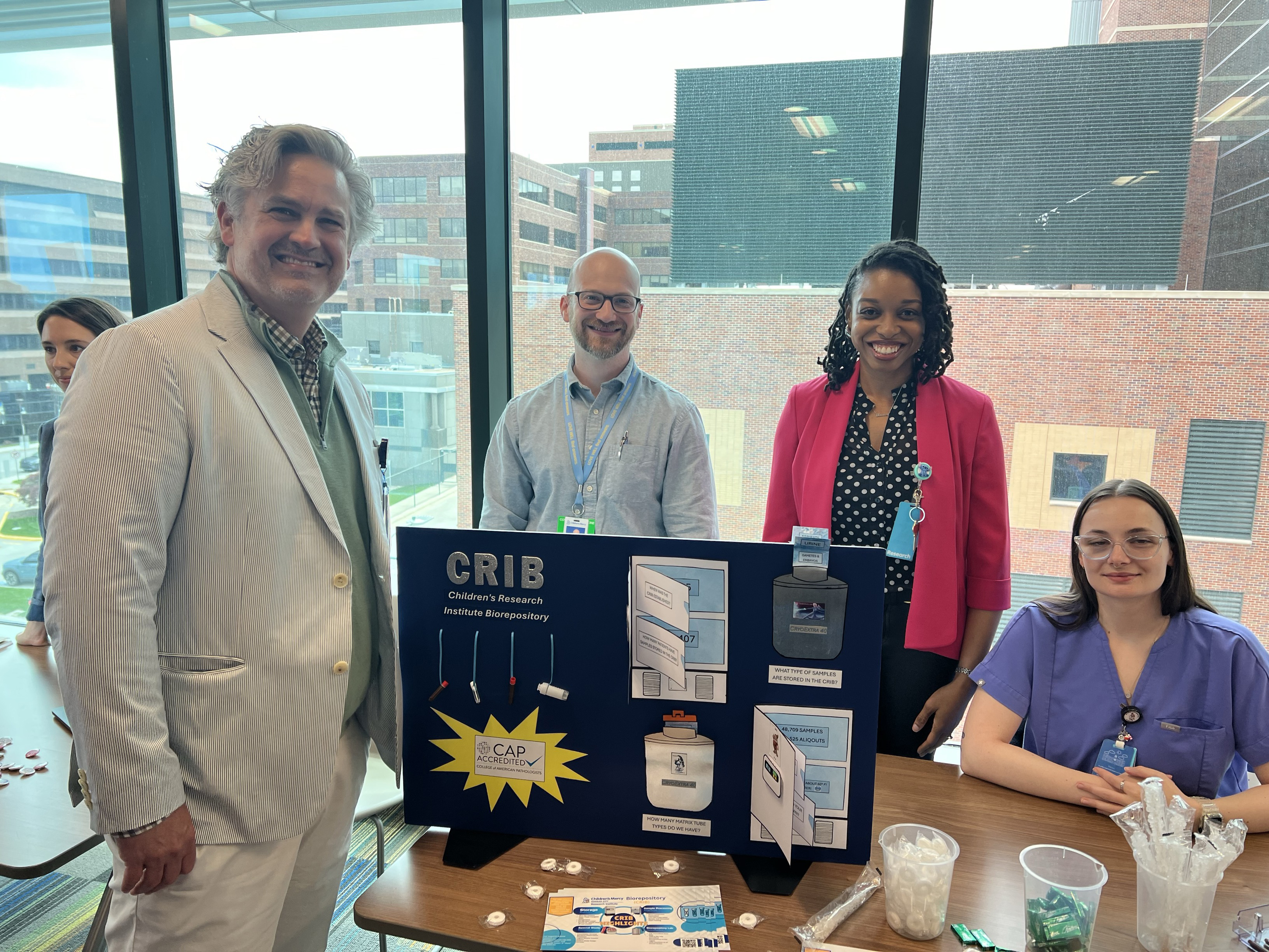 Two men and two women pose together. 3 stand while 1 sits. At the table they pose around is a sign that says CRIB, Children's Research Institute Biorepository.