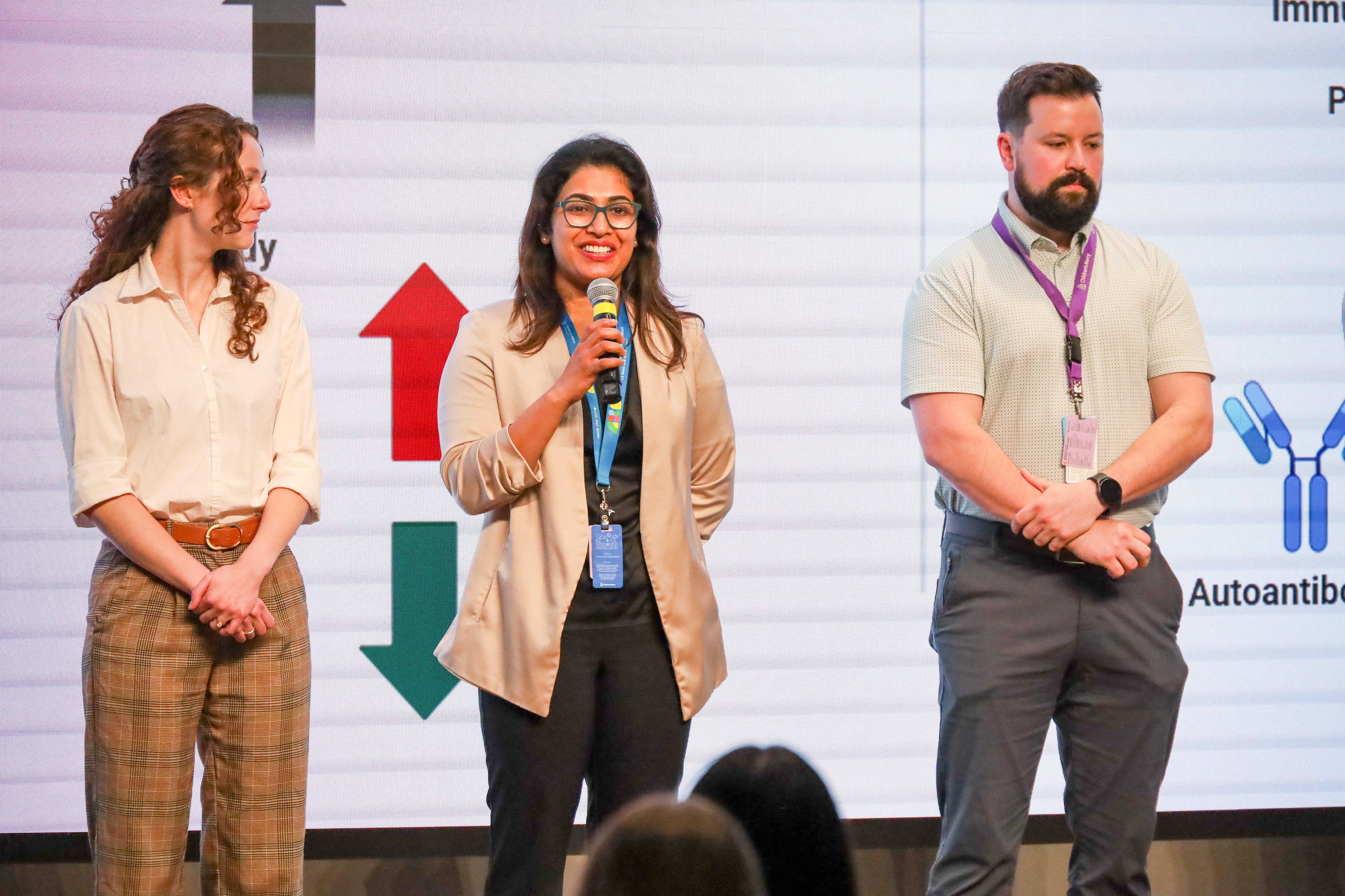 3 people stand on stage, 2 women and 1 man. The woman in the middle holds a microphone, speaking.