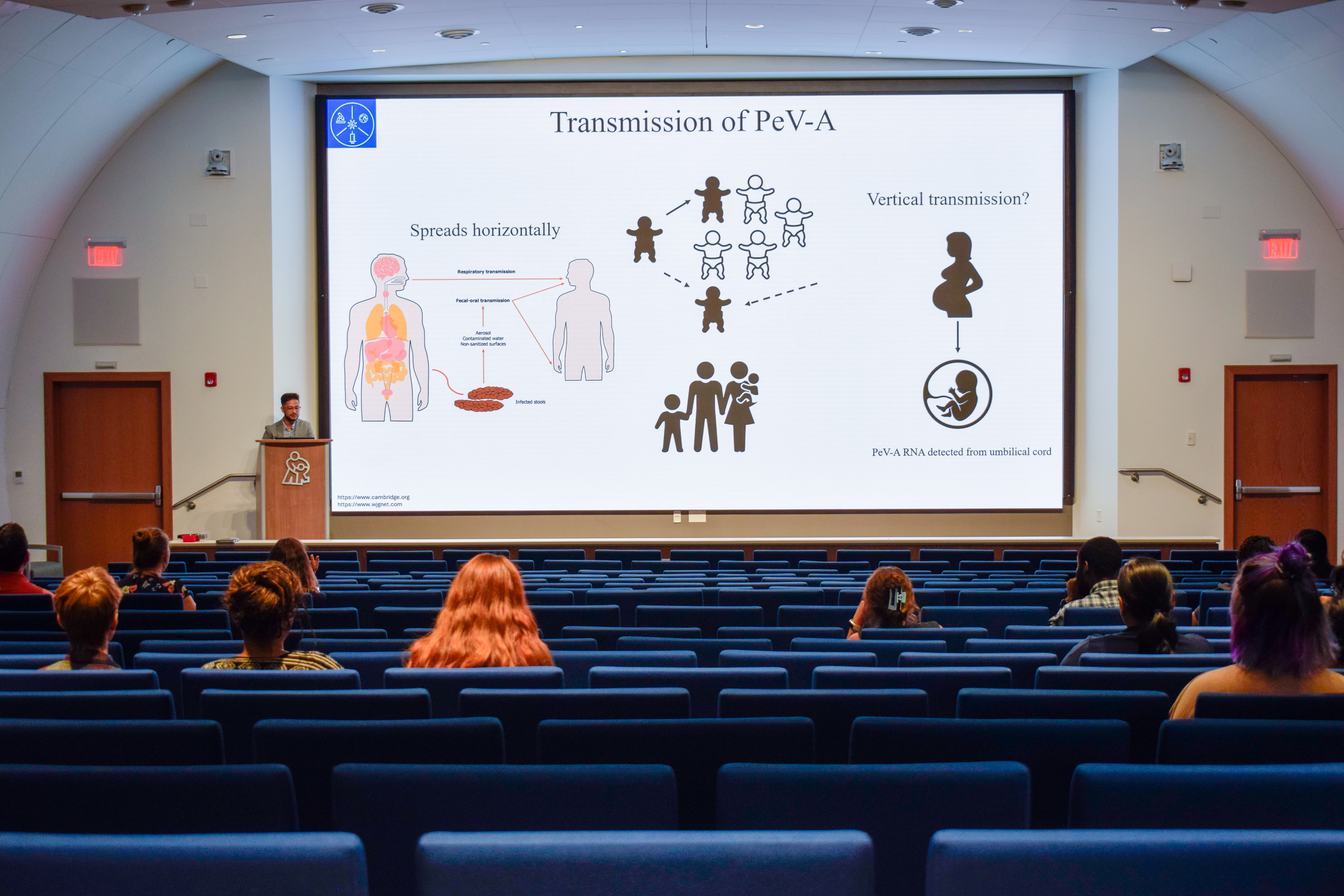 A man stands at a lectern presenting to an auditorium of people. On the screen behind him is a slide that's titled Transmission of PeV-A.