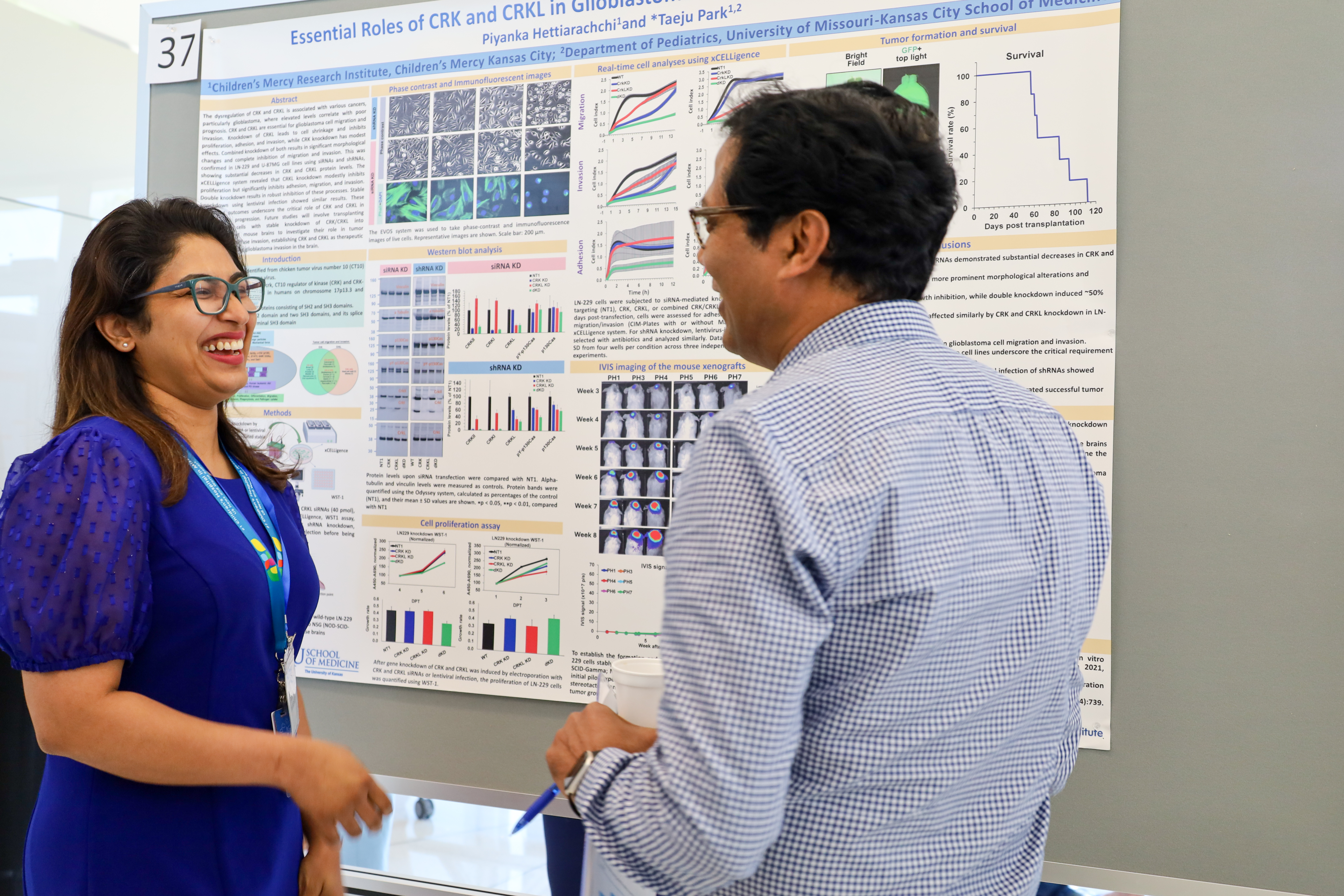 A man and a woman stand in front of a research poster. Both are laughing. The woman is wearing a blue dress and the man is wearing a plaid, button-up shirt.