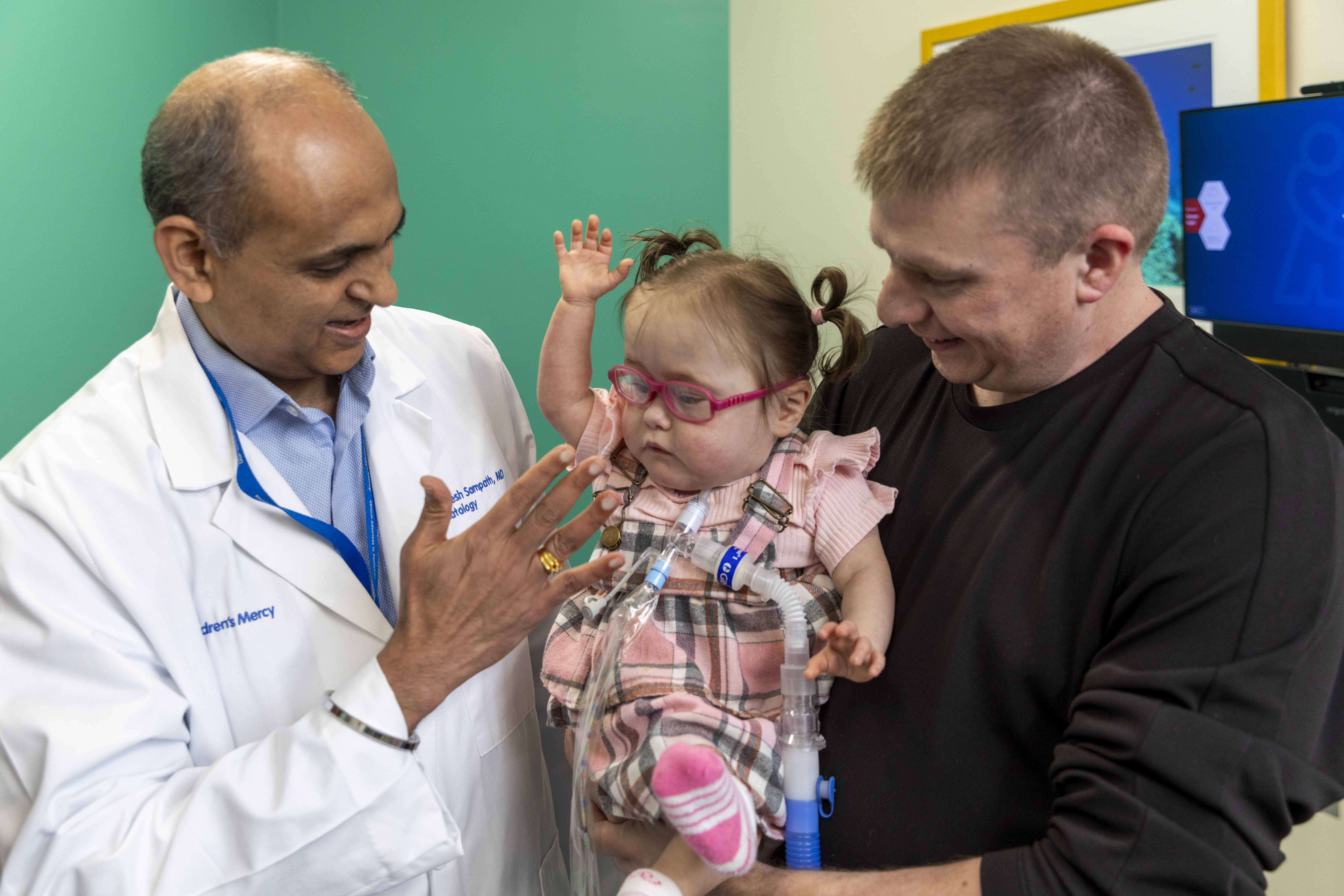 Dr. Sampath stands with a father. Together they hold a young girl with a tracheostomy tube and pink glasses.