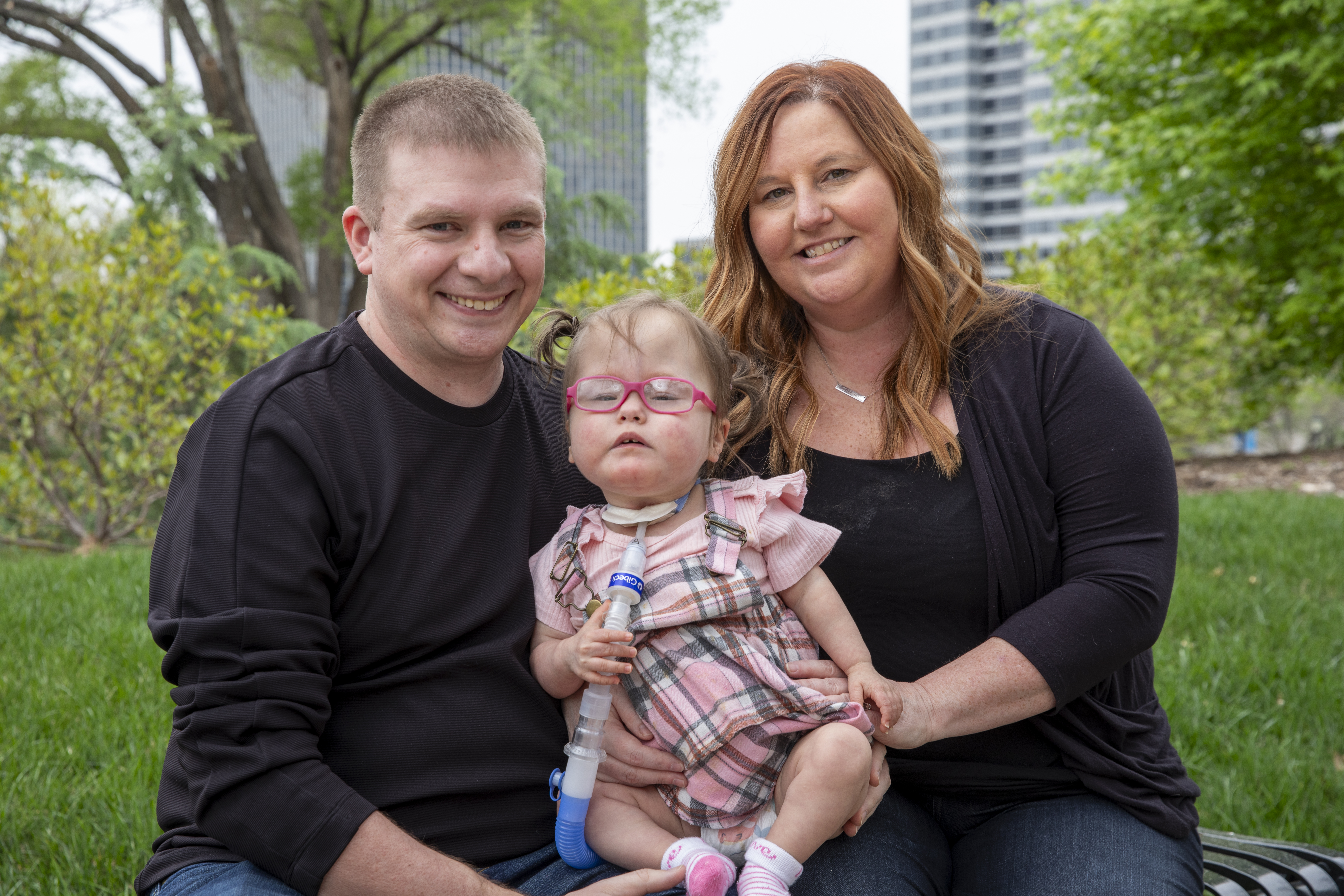 A mother and father sit outside with their young daughter. She has a tracheostomy tube and pink glasses.