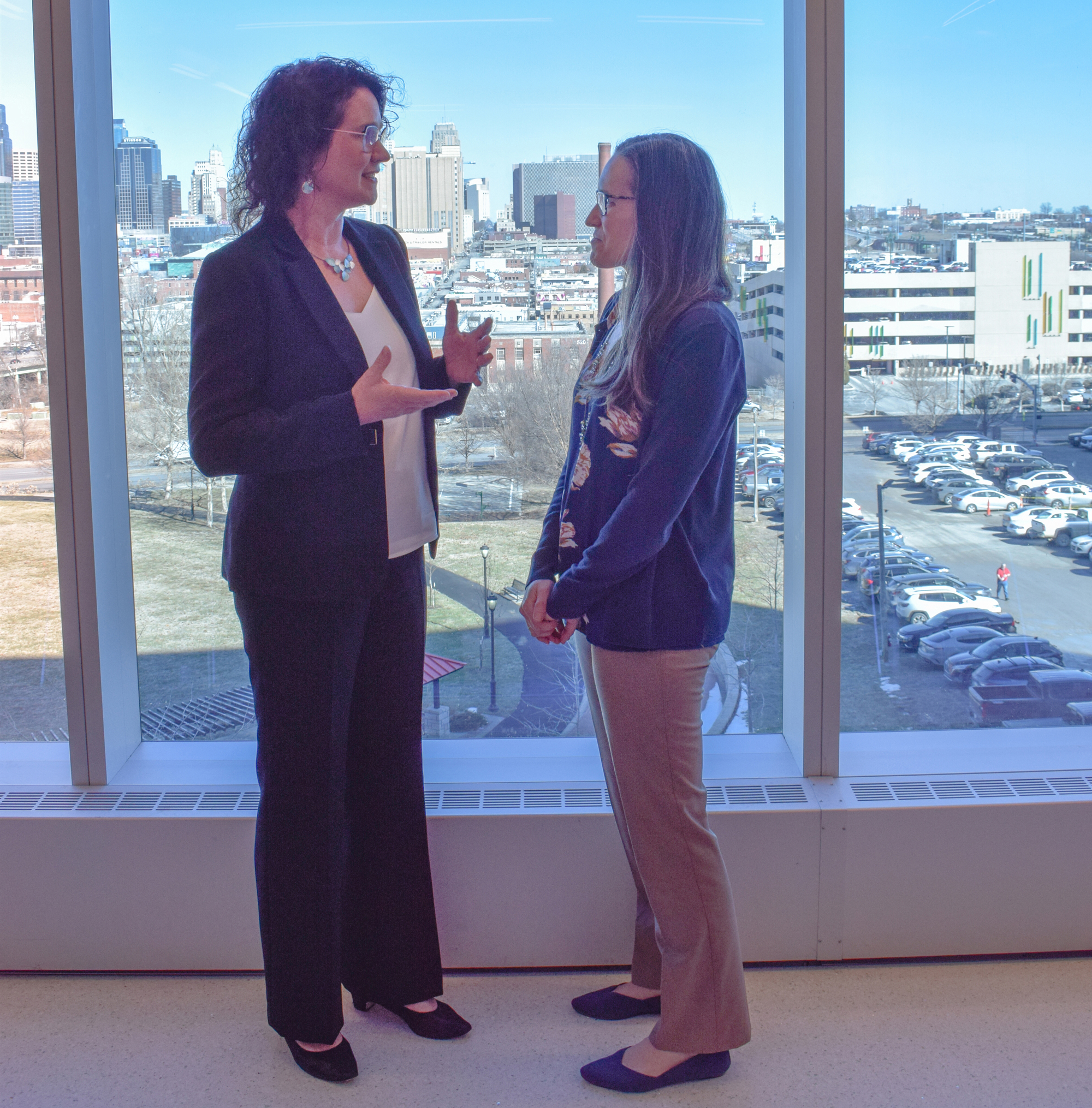 Two women stand by a large glass window, talking. The woman on the left is motioning with her hands. Behind them, outside, is a downtown cityscape.