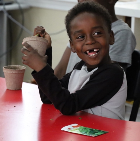 A young boy sits at a table, smiling. He is holding a pot with a plant.
