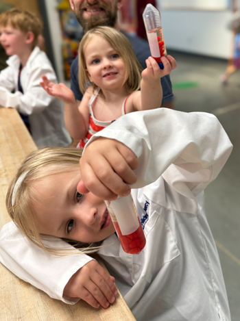 Young girls sit at a table wearing lab coats and holding large tubes of a liquid mixture that is clear and red.