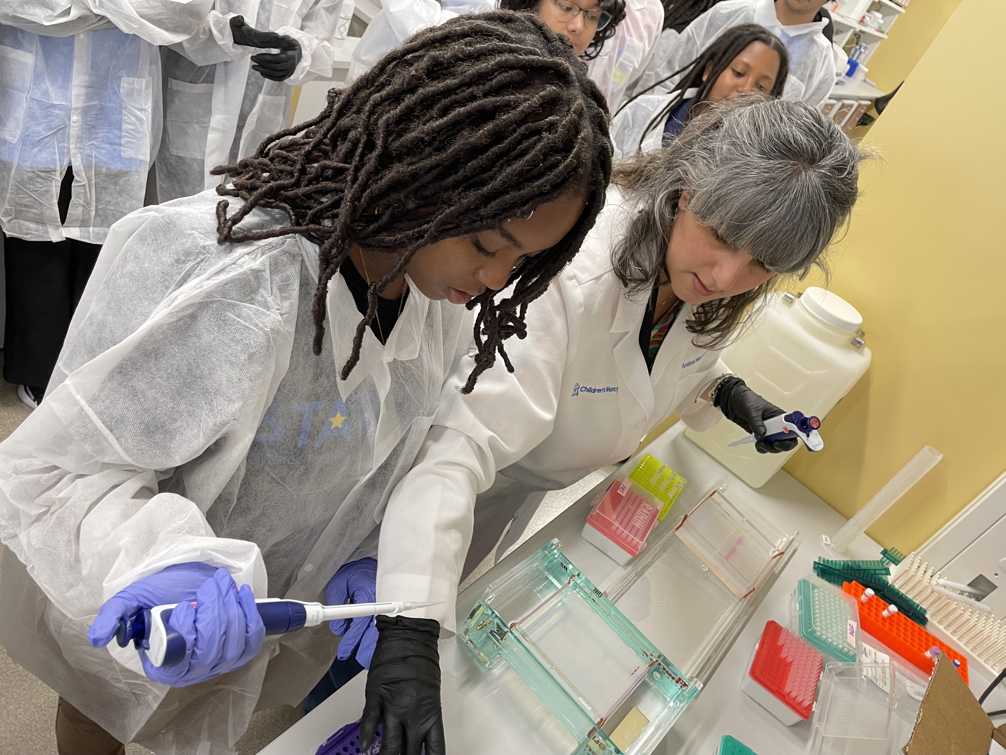 A woman and several teenagers stand in a lab. They are wearing lab coats and she is showing them how to use a piece of lab equipment.
