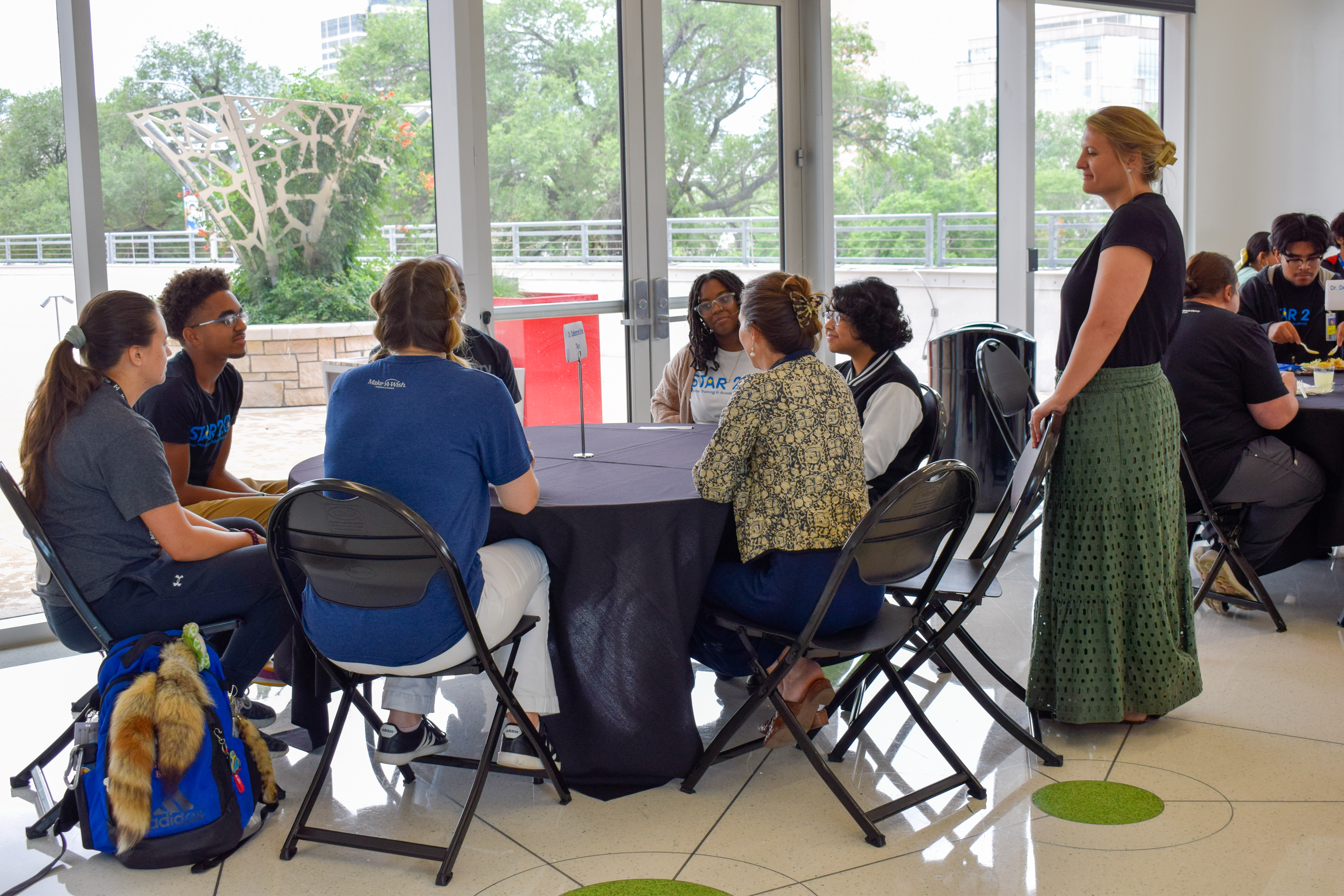 A group of teens sit at a round table. A woman with blonde hair, a black shirt, and a green long skirt stands next to the table.
