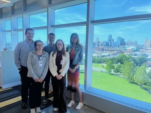 Dr. Stancil stands with her research team, made up of 2 men and 2 women. A large window is behind them and it is sunny out. A cityscape is visible in the background.