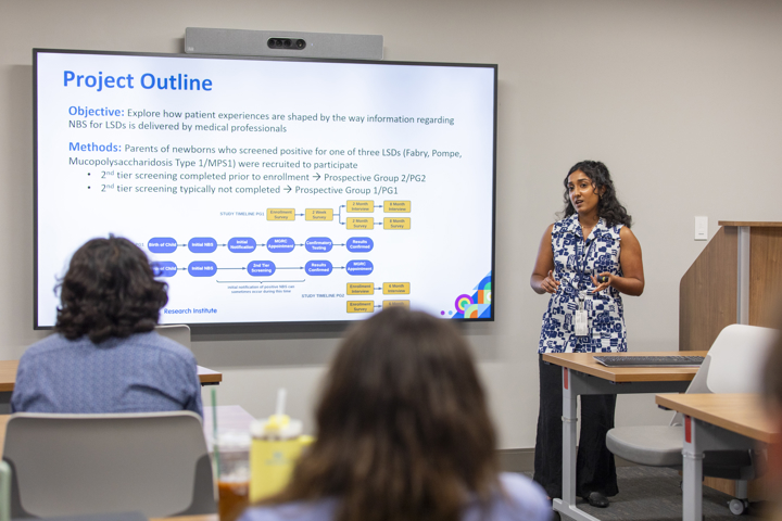 A woman stands in front of people, presenting. She has black hair. Behind her on a screen on the wall is a slide that says Project Outline and outlines objectives and methods.