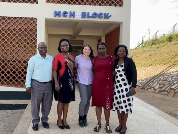 5 people (one man and 4 women) stand together outside. Behind them is a building that says MCH Block. The photo is taken in Uganda.