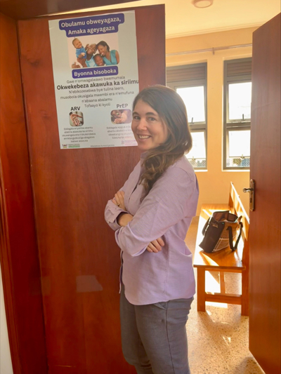 A woman stands inside next to a poster promoting new HIV testing communication strategy. The poster features the availability or pre-exposure prophylaxis and themes of healthy relationships and childbearing. 