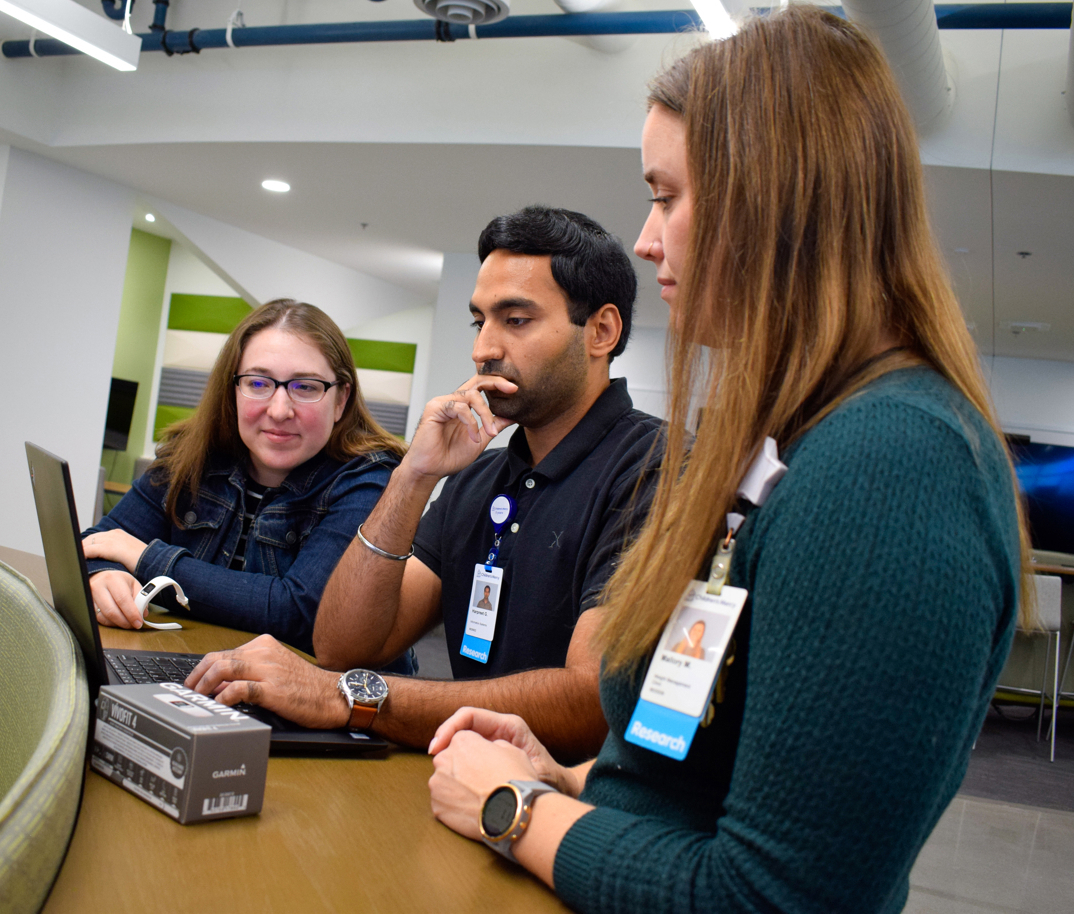A woman, man, and another woman stand together looking at an open laptop. A box for a fitness tracking watch sits on the table and the woman on the left holds the fitness tracker.