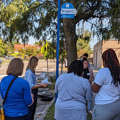 A group of people stand outside at a bus stop. They are being given instructions and holding papers.