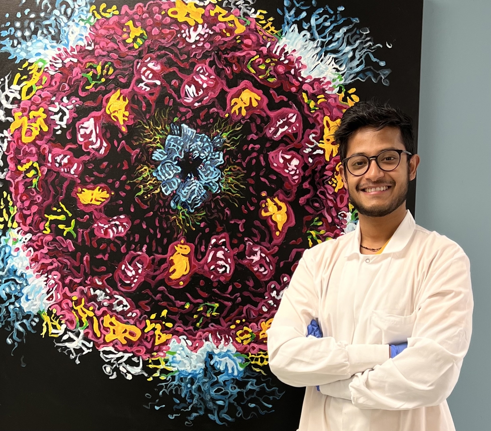 Debarpan Dhar. He is wearing a white lab coat and stands in front of a colorful painting.