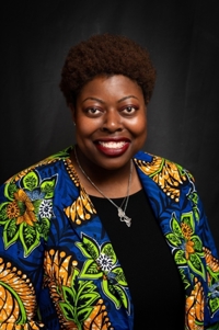 Lynette Lartey. She has black short, curly hair. Her jacket is blue with colorful plants on it. She's also wearing a black shirt and a necklace.