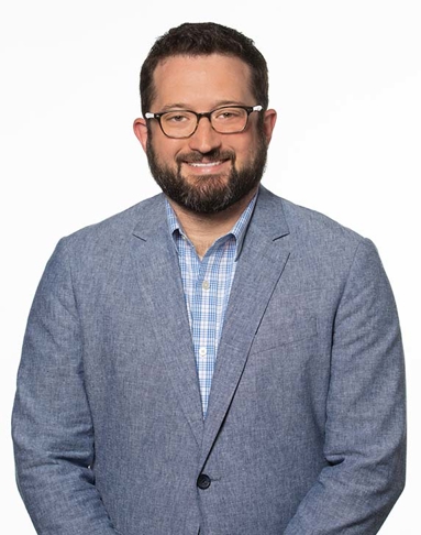 Pete Muenks. He has dark hair and is wearing glasses. He is wearing a denim-looking blazer with a plaid shirt underneath.