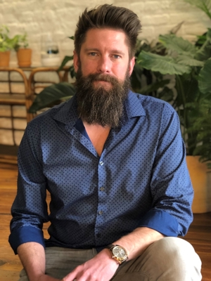 Nik Schuetz, vertical photo. He has brown hair and a full beard. He is wearing a blue button-up shirt and khakis. He sits, posing, with plants in the background.