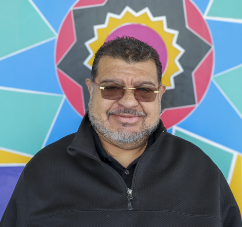 Luis Sainz. He has dark short hair and wears sunglasses. He stands in front of a colorful sun and star mural.