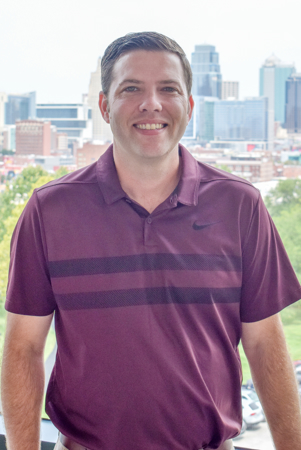 Sean Bender. He has brown short hair and wears a maroon polo. He stands in front of a window with a downtown behind him.