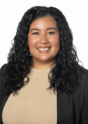 A portrait of Selina Tucker. She has long black curly hair and is wearing a tan shirt with a black blazer over it.