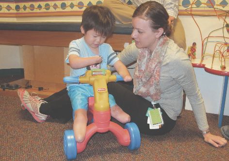 Therapist sitting beside a child riding a toy tricycle during a developmental or rehabilitation therapy activity in a pediatric clinic
