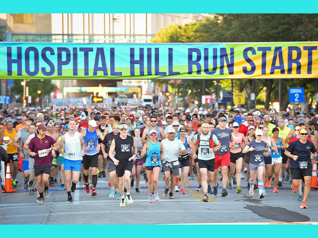 Large group of runners crossing the start line of the Hospital Hill Run road race beneath a banner reading 'Hospital Hill Run Start.'