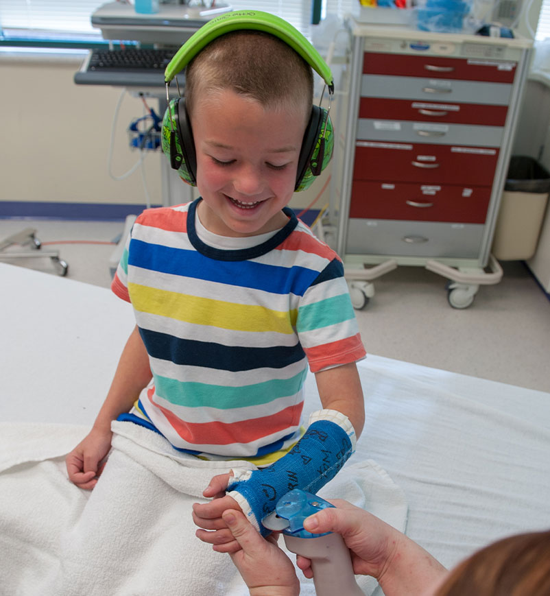 image of smiling child sitting on a bed watching the clinician start to remove the cast